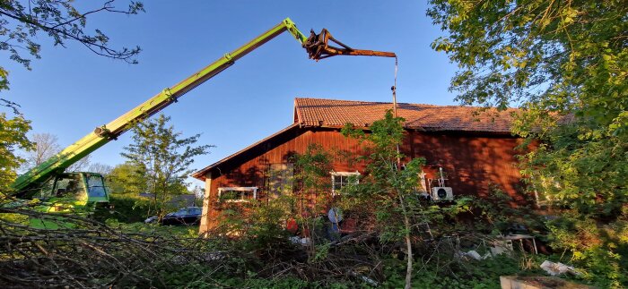 Kran lyfter en skorsten från ett rött garage omgivet av grönska och nedklippta buskar.