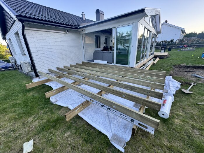 Pågående altanbygge med träbjälkar monterade på stödblock vid en husvägg och en uterumskonstruktion i bakgrunden.