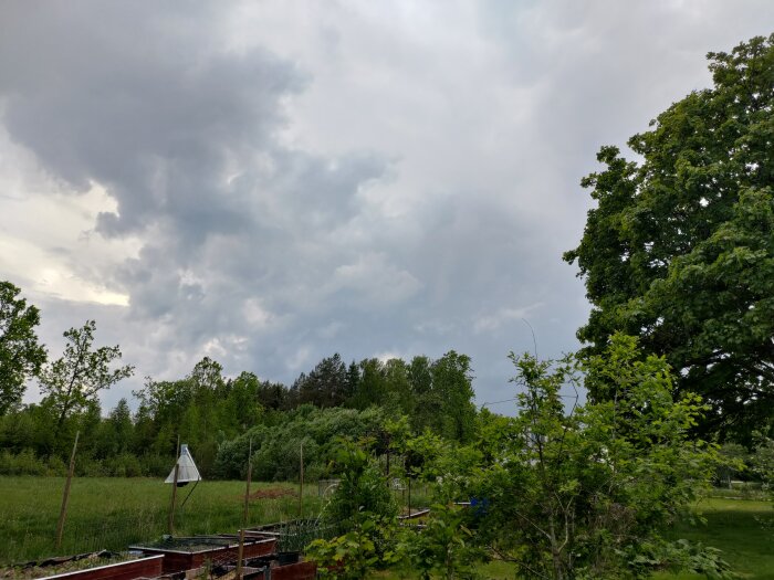 Mörka regnmoln på himlen över en grön trädgård, regnet syns i fjärran men inte närmare.
