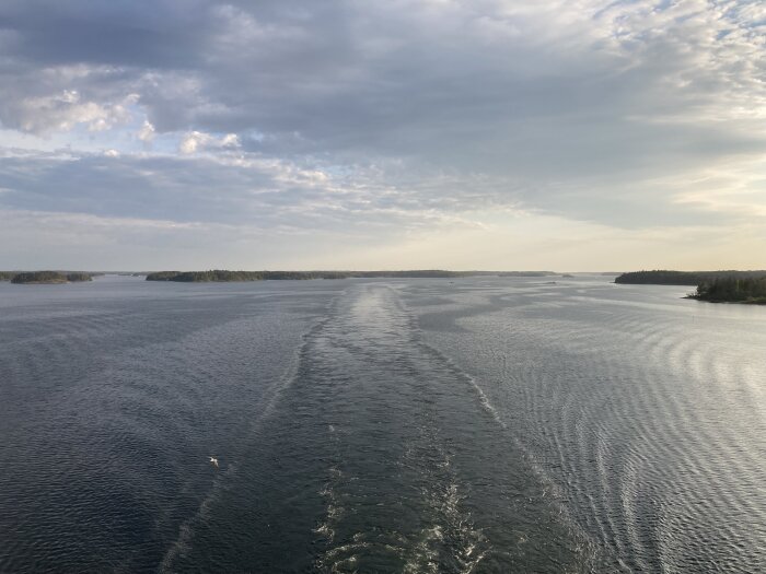 Båtvågor på ett stilla hav omgivet av skogklädda öar under en molnig himmel.