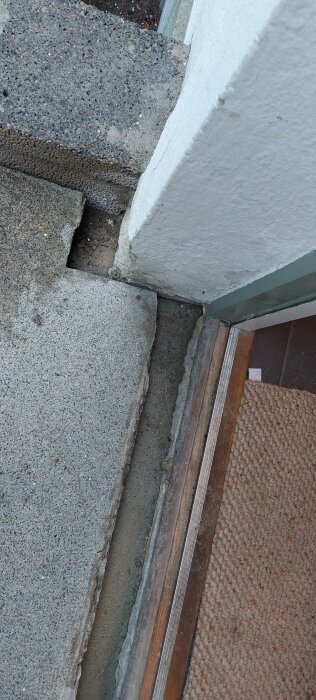 Bild på ett glapp mellan fasad och trappsteg vid en husentré, med synlig metalltröskel och kantsten.
