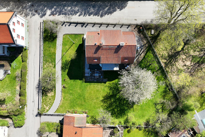 Flygbild av ett litet hus med tegeltak till höger omgivet av grönska och blommande träd.