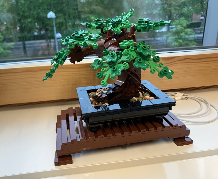 Ett legobyggt bonsai-träd med gröna löv på en brun trädstam, placerat på en fönsterbräda inomhus.