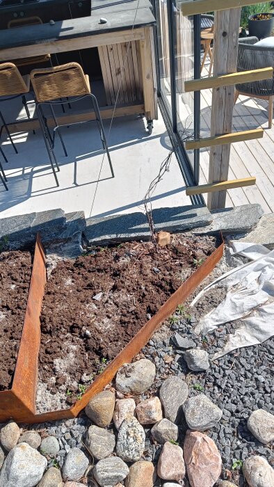 Upphöjd bädd med plåtkant och stenmur, jord och en nyligen planterad planta, uteplats i bakgrunden.