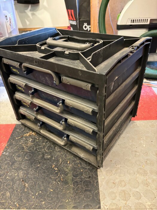 Dammig svart verktygslåda med flera lådor i ett garage, oanvänd och till salu.