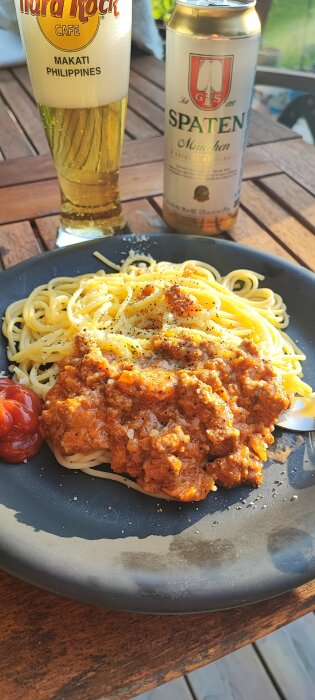 Tallrik med spagetti bolognese och kryddor, ölglas och burk på träbord utomhus.