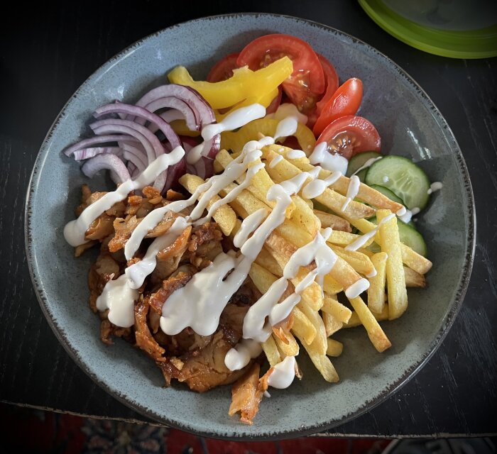 Kebabtallrik med kyckling, pommes, färska grönsaker och vit sås drizzlad över, serverad på en grå tallrik.