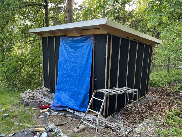 Ett halvfärdigt bygge av en svart uthus med blå presenning och byggmaterial runtomkring i skogsmiljö.