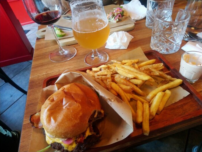 Baconburgare med pommes och chillimayo på serveringsbricka, öl i glas och rödvin, med suddig bakgrund.