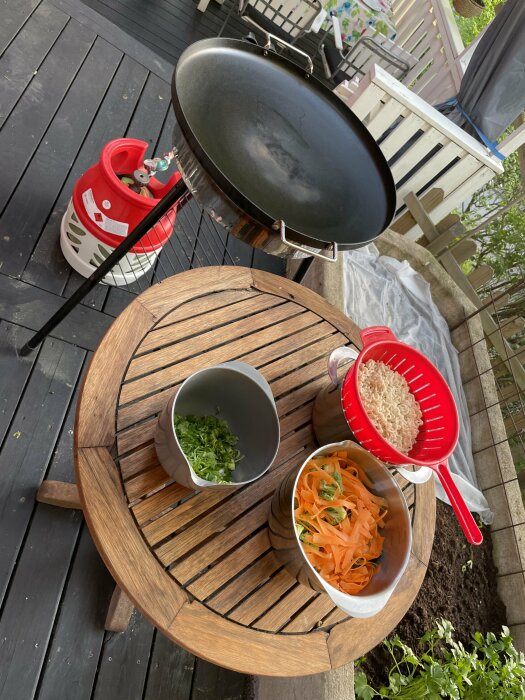 Grill och matförberedelser på en terass med skivade grönsaker, nudlar och gasolbehållare.