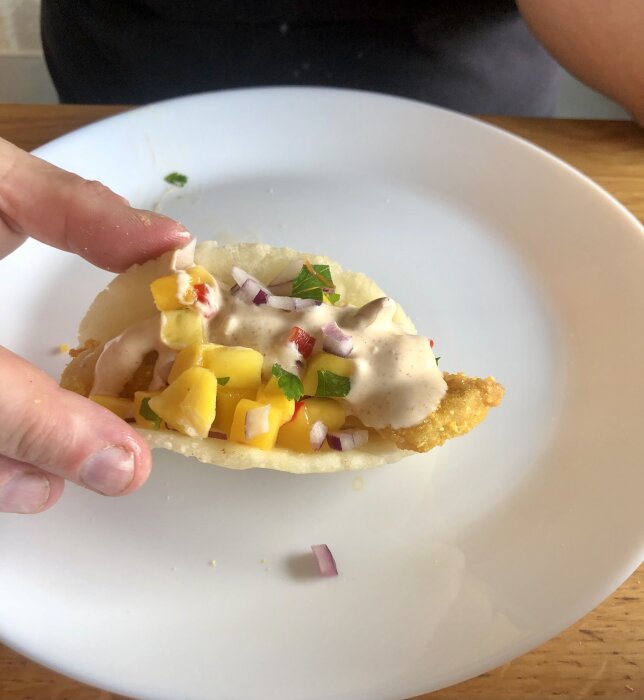 En hand håller en fisktaco med mango, lök och sås på en vit tallrik.