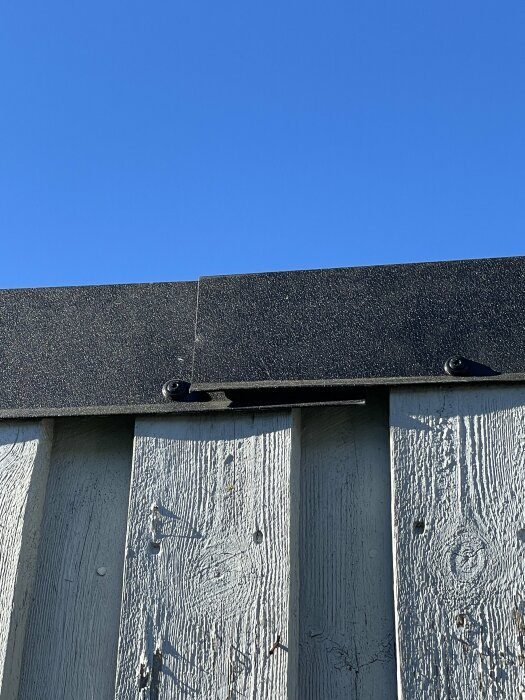 Detaljbild på en vit trädvägg med en svart takfotpanel och synliga skruvar mot en klarblå himmel.