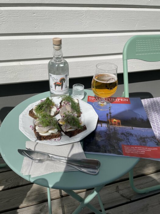 Liten utomhuslunch med smörgåsar toppade med sillinlägg, dill och lök, en flaska sprit, ett ölglas och en tidning på ett turkost cafébord.