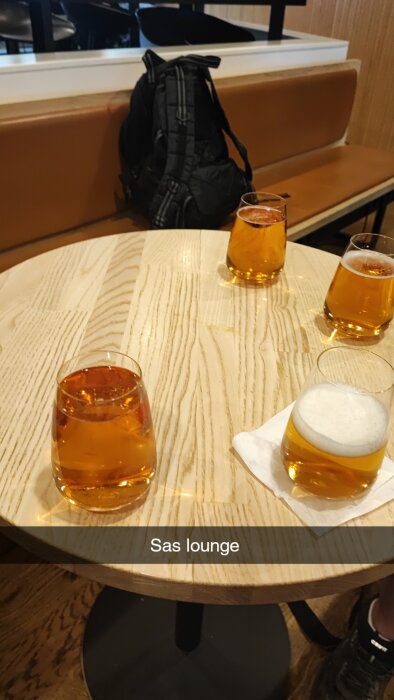 Tre ölglas med gyllene dryck på ett träbord i en lounge, en ryggsäck i bakgrunden.