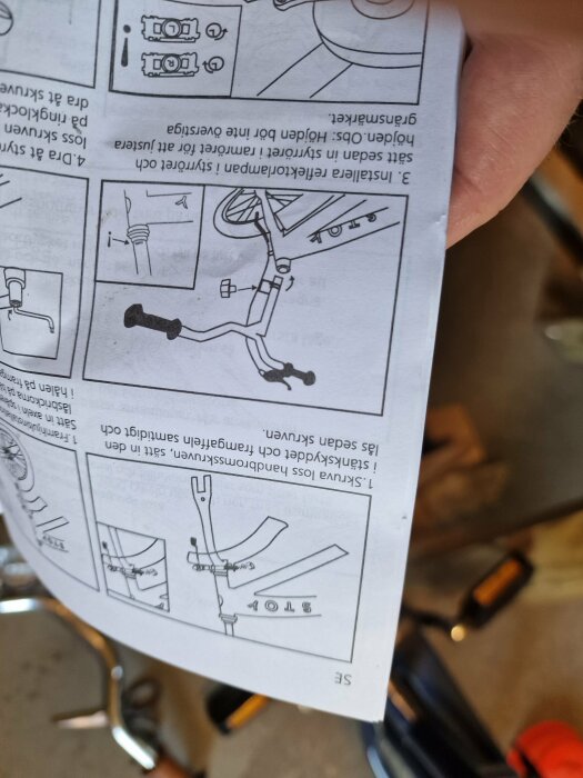 Upp och nervänd bild av en hand som håller en manual med instruktioner för montering av cykelstyre.