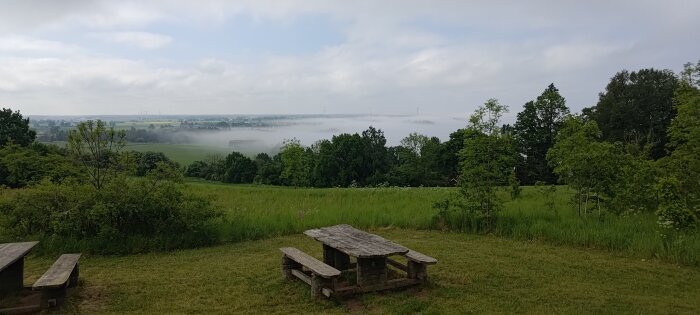 Utsikt över en dimtäckt landsbygd med två träpicnicbord i förgrunden.