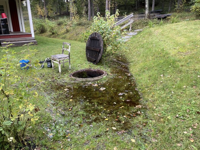 Gammal brunn med vattensamling i gräsmattan, pump och trädgårdsstol intill, på landet.