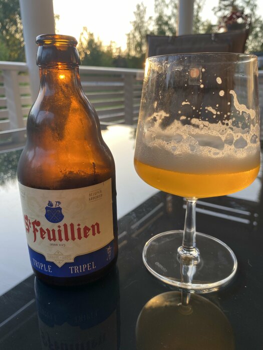 En flaska belgisk öl av märket St. Feuillien Tripel bredvid ett halvfullt glas på ett bord utomhus.