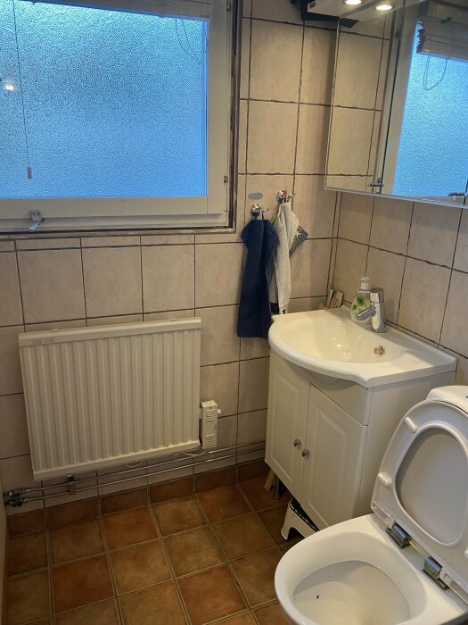 Badrum före renovering med kaklat golv, vägg, WC, handfat och radiator framför ett frostat fönster.