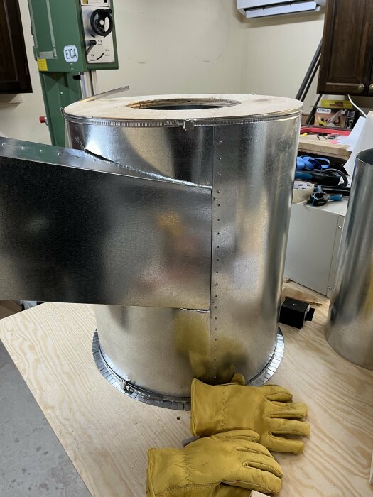 Metallcylinder med popnitade fästen och borrhål på en verkstadsbänk med gula arbetshandskar.