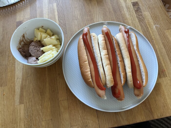 Tre varmkorvar med bröd och ketchup, skål med glass och ananasbitar på en träbordsskiva.