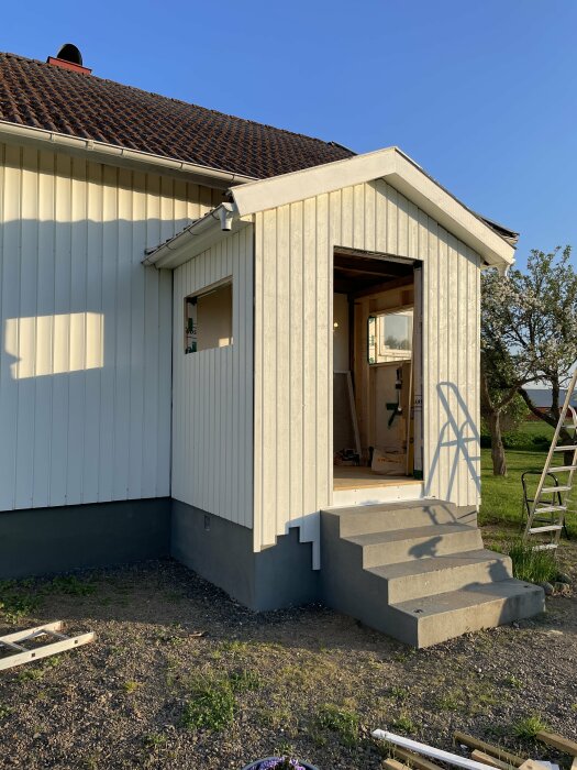 Nyinstallerad vertikal träpanel på en verandas vägg vid ett hus med tegeltak och trästege i bakgrunden.