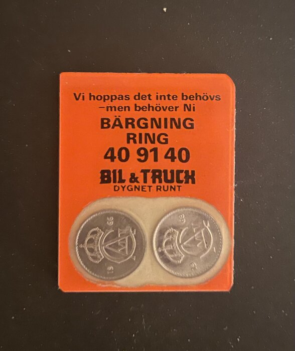 Bild på en orange magnet med text "Bärgning Ring 409140 Bil & Truck Dygnet Runt" och två mynt.