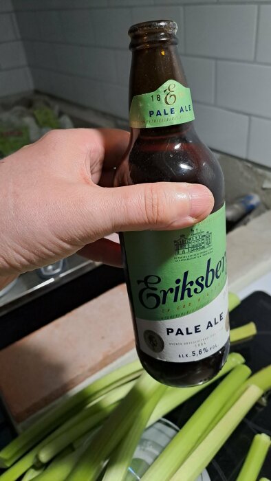 Hand håller en flaska Eriksberg Pale Ale framför hackad selleri på en köksbänk.