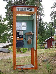 Gammal telefonkiosk i trä med öppen dörr och en röd stuga i bakgrunden.