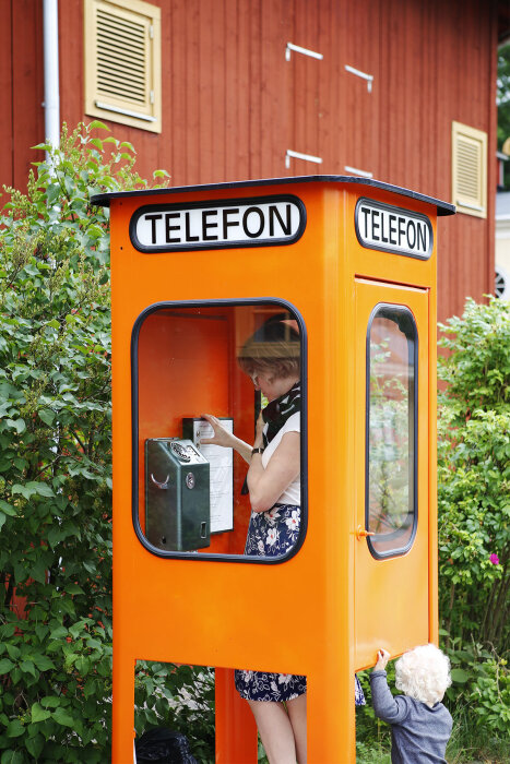 Orange telefonkiosk med en person inuti som använder en telefon och ett barn utanför.