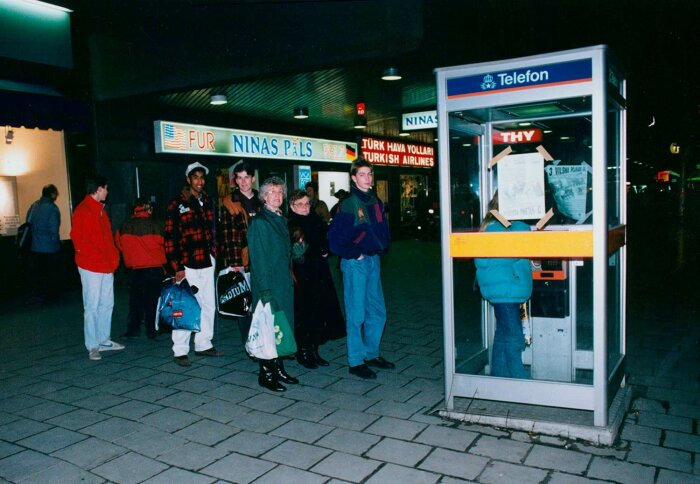 Människor köar utanför en modern telefonkiosk på en välbesökt plats i stadsmiljö.