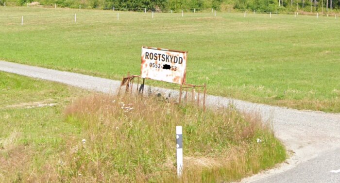 Rostig skylt med texten "ROSTSKYDD" vid en vägkant, omgiven av grönt gräs och vildblommor.
