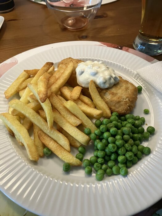 Tallrik med fish and chips, créme fraîche-röra, och gröna ärtor, serveras med ett glas öl i bakgrunden.