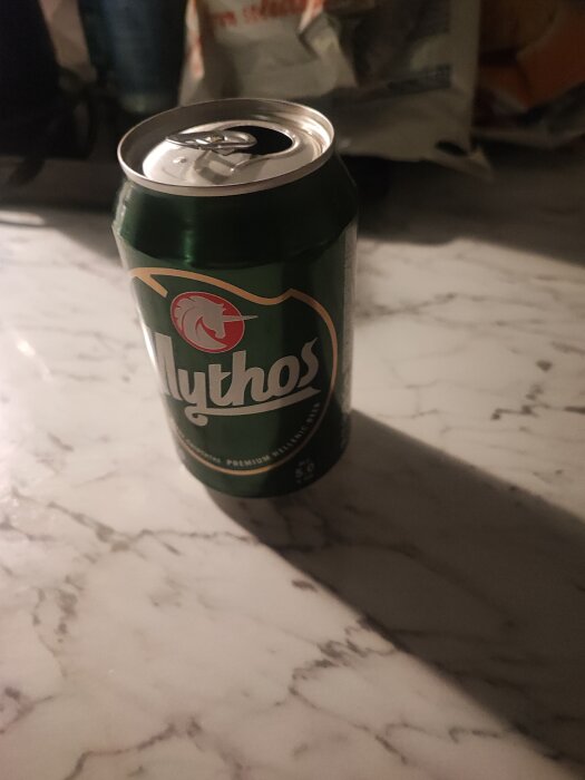 Öppnad burk av Mythos öl på ett marmorbord, med suddig bakgrund.