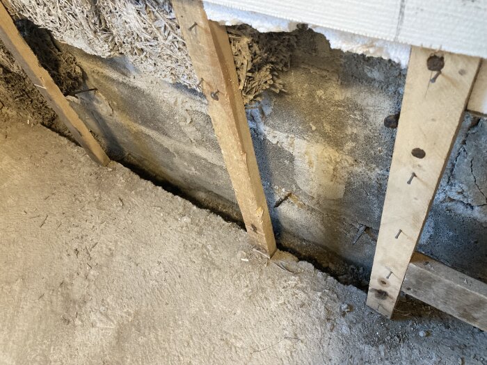 Delvis rivet golv och vägg i rum med synlig fuktskada och träullit isolering, träreglar och betongfundament.