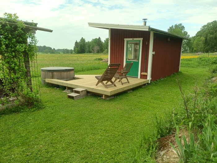 Trädäck vid röd stuga med två trädgårdsstolar och ett badkar utomhus, omgivet av grönt fält.