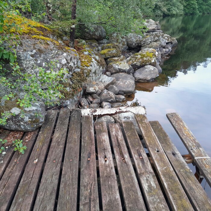 Gammal träbrygga med skadade plankor vid insjö, omgiven av stenar och vegetation.