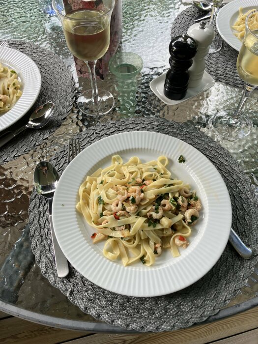 Pasta med rÀkor, fÀnkÄl, chili och persilja pÄ ett vitt fat med ett glas vitt vin och kvarnar.