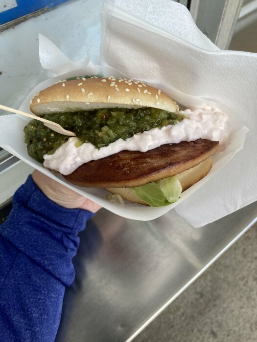 Hand hÄller en parisare med rÀksallad i bröd med sesamfrön, inlindad i vitt papper.