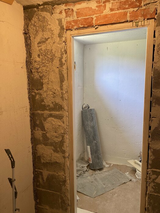 Delvis putsad vägg med exponerade tegelstenar och en öppen dörr till ett rum med skyddspapper på golvet.