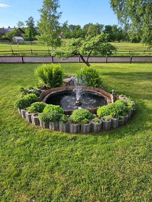 Trädgårdsfontän med gröna växter och krukor mot lantlig bakgrund.