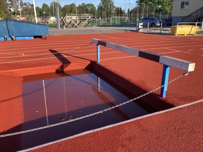 Gymnastikbänk på röd löparbana med spegling i vattenpöl, tidig morgon eller sen eftermiddag.