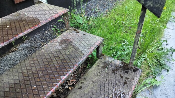 Rostig, metallgallerklädd ståltrapp utomhus med synlig slitage och växtlighet runt omkring.