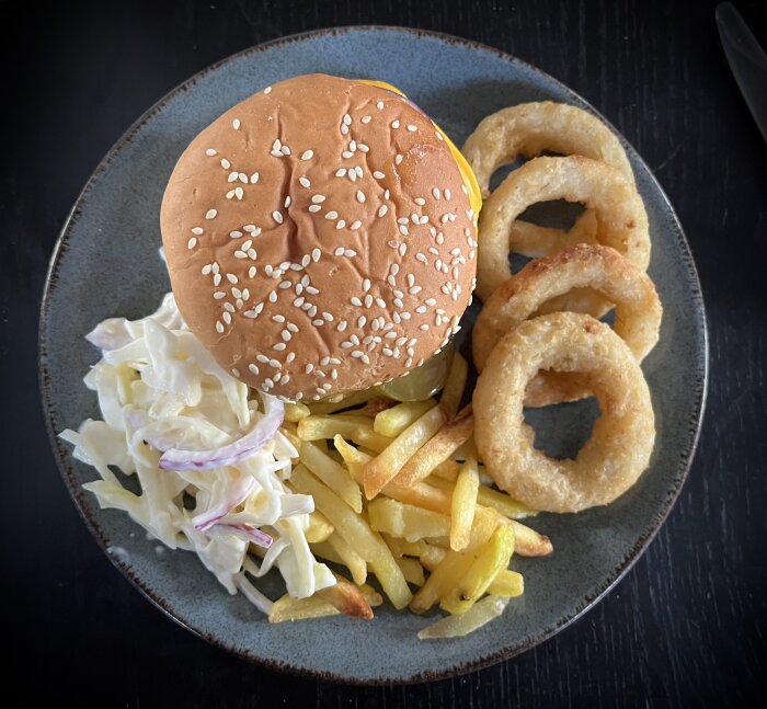 Hamburgare med sesamfrön pÄ brödet, serverad med pommes frites, lökringar och coleslaw pÄ en tallrik.