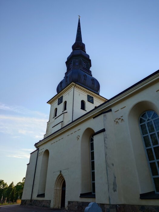 Kyrkobyggnad med lutande svart spira mot blå himmel, vita väggar och fönster med bågar.