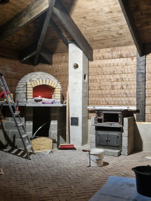 Byggarbetsplats med vedeldad pizzaugn, stenkakelugn och träbeklädnad under konstruktion.