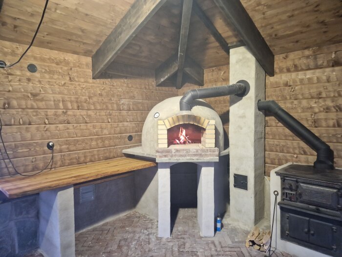 Inbyggd vedeldad pizzaugn i rustik stil med öppen eld, intill en klassisk vedspis i ett träbeklätt rum.