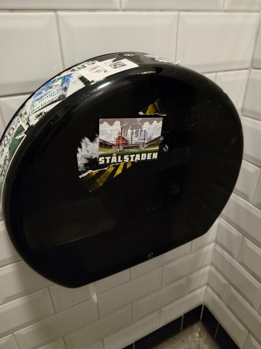 Svart toalettpappershållare med klistermärken och text "STÅLSTADEN" monterad på vit kakelvägg.