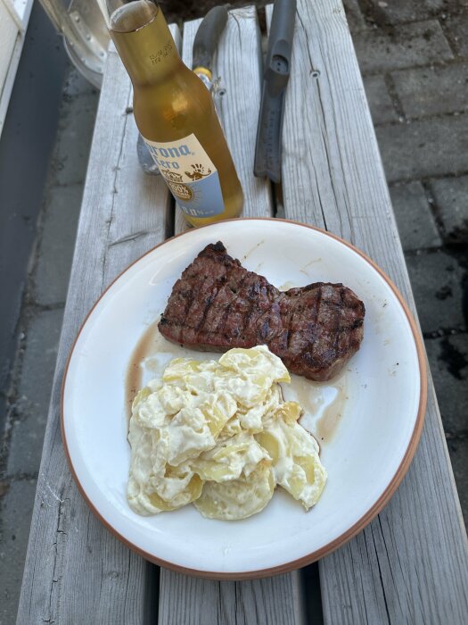 Grillad biff med potatissallad pÄ tallrik bredvid ölflaska och grillverktyg pÄ trÀbord.