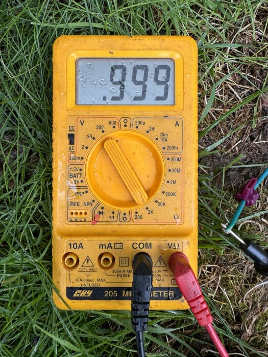 Multimeter som visar 999 ohm, ligger på gräsmark med kopplade testledningar.